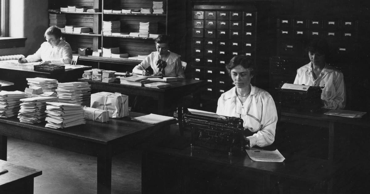 A black and white photo of four women sitting at desks with typewriters, stacks of papers, and card catalogs. 
