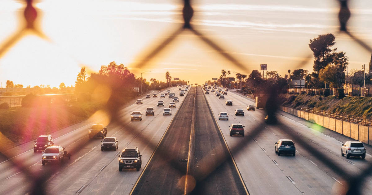 View of a freeway seen through a chain-link fence at sunset. 