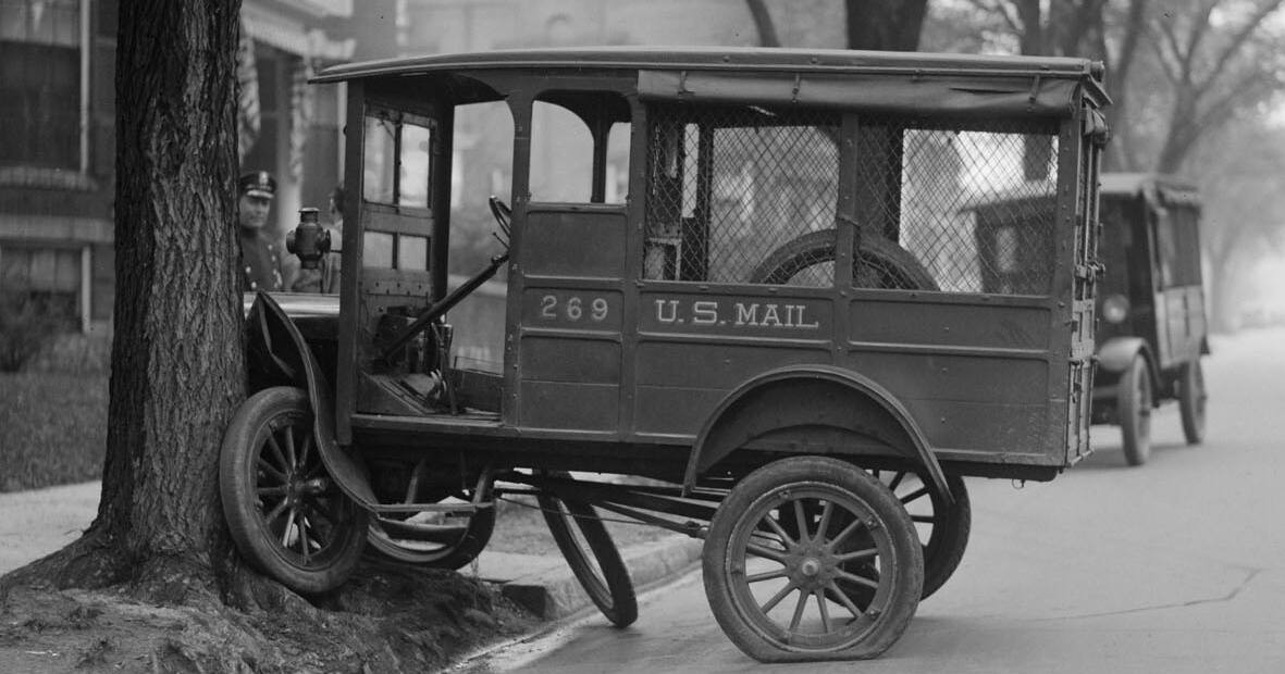 A black and white photo from 1927 of an old USPS truck crashed into a tree at the side of the road. 