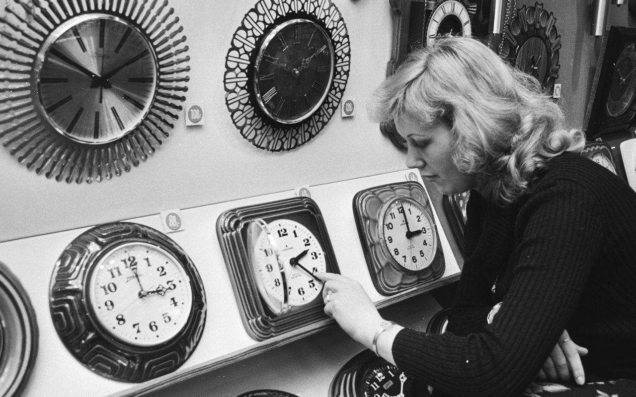 A blonde woman adjusts the time on a row of clocks at a store. 
