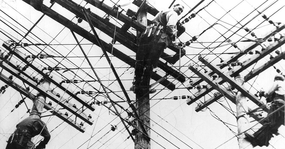 A black and white photo of three linesmen repairing a tangle of overhead wires. 