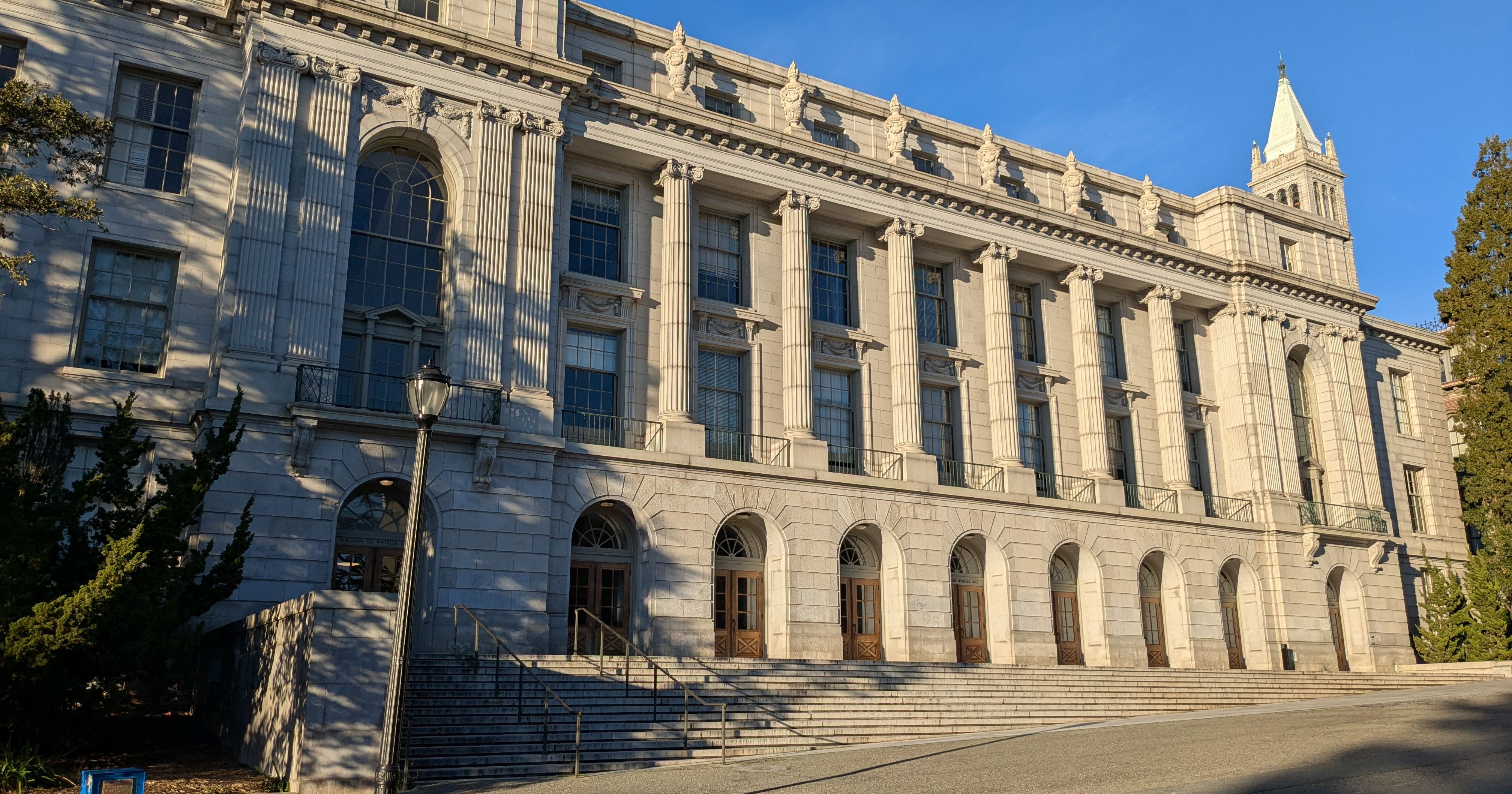 The grand, neoclassical facade of Wheeler Hall on the UC Berkeley campus, viewed from a low angle in the late afternoon. The building's stone columns and wide steps are highlighted by strong sunlight and shadow, with the Campanile visible in the background against a clear blue sky.