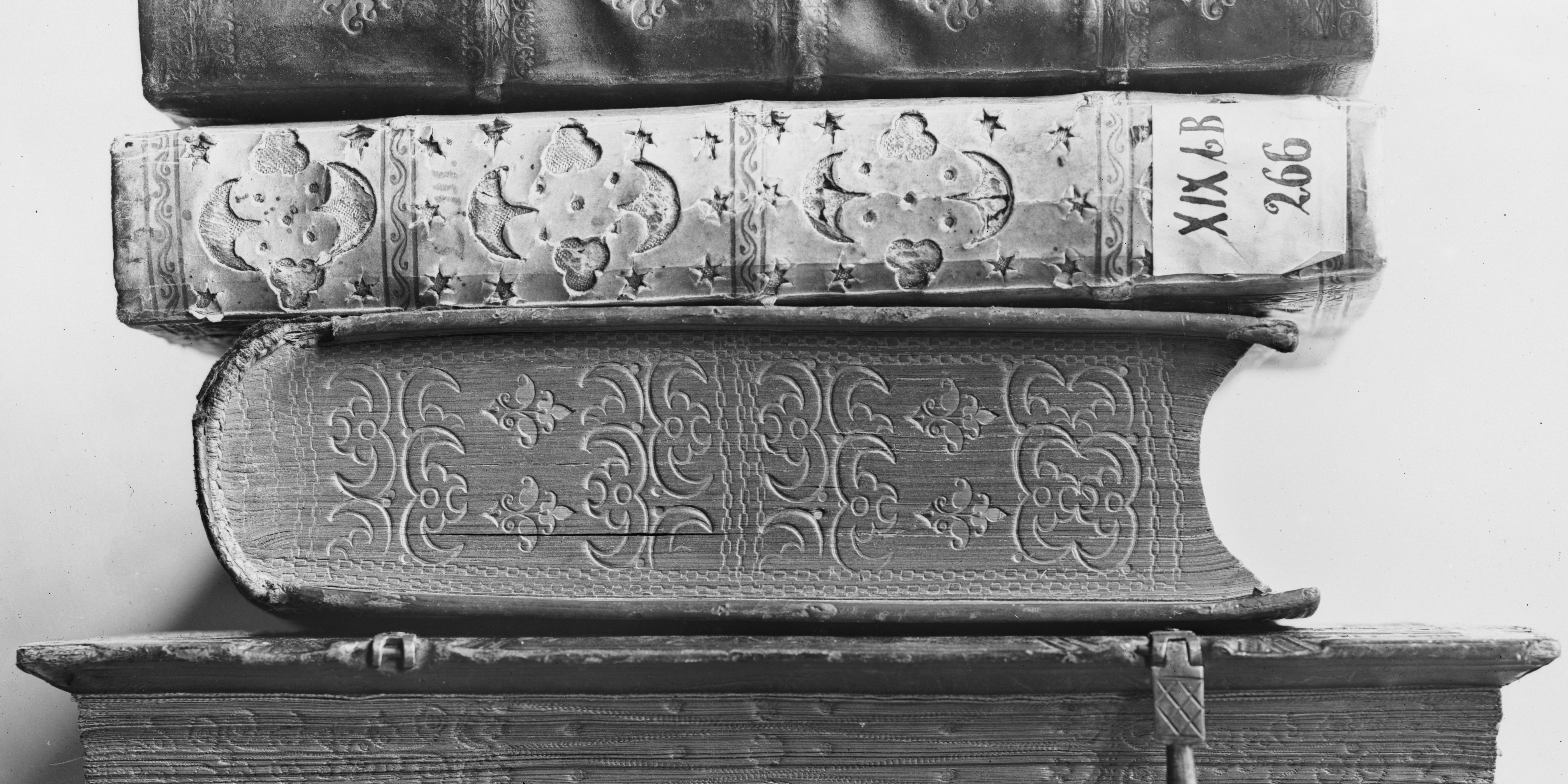 Black-and-white photograph of a stack of four old, thick books seen from the side, showing their spines and page edges. The books are richly decorated with embossed floral and geometric patterns, worn leather bindings, and visible aging. One book has a small handwritten label on its spine, and another is secured with a metal clasp. The overall appearance suggests antique or historical volumes. 
