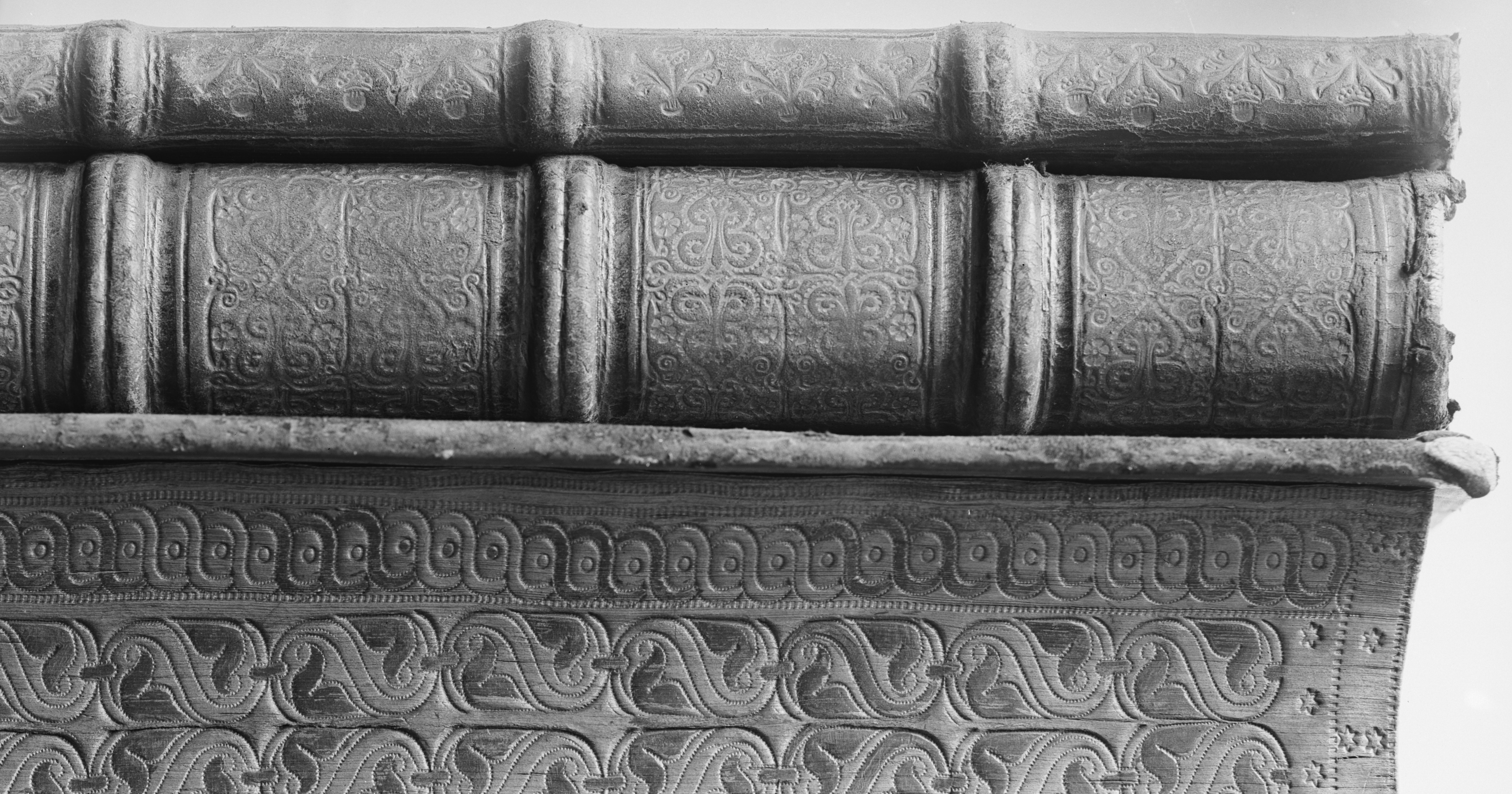 Black-and-white close-up of three horizontally stacked antique books viewed from the side. The worn leather bindings feature raised bands and elaborate embossed patterns, including repeating scrolls, floral motifs, and geometric borders. Frayed edges and surface wear emphasize the age and craftsmanship of the volumes. 