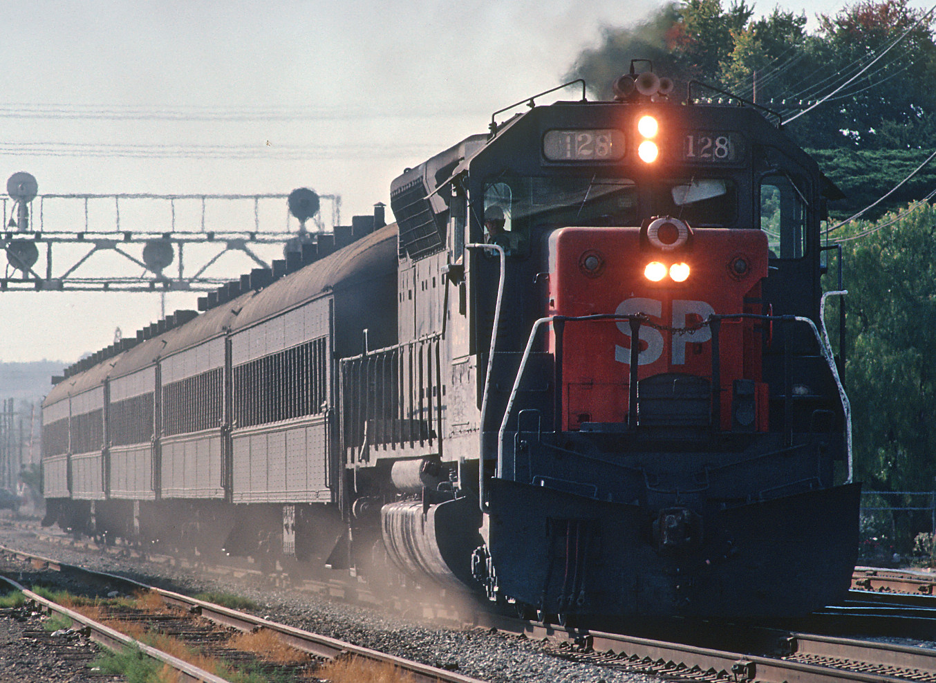 A Southern Pacific Railroad commuter train in Redwood City, circa August 1980. The train is grey with a bright red square painted on the front containing the letters 'SP' in white. 