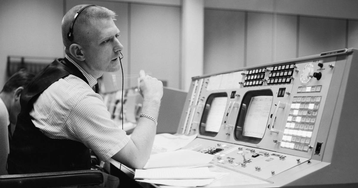 Gene Kranz sits at a console in the NASA Mission Operations Control Room. He is wearing a single ear headset and flipping a pencil back and forth. 