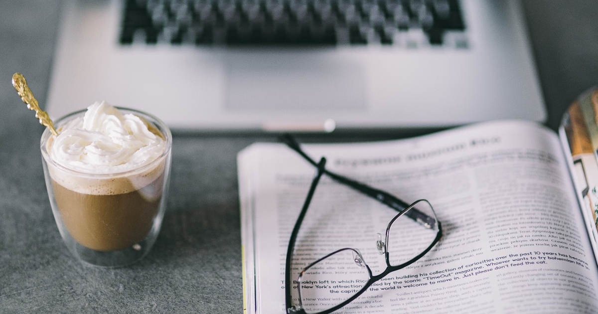 A coffee, reading glasses, and a book sit on a table in front of a MacBook.