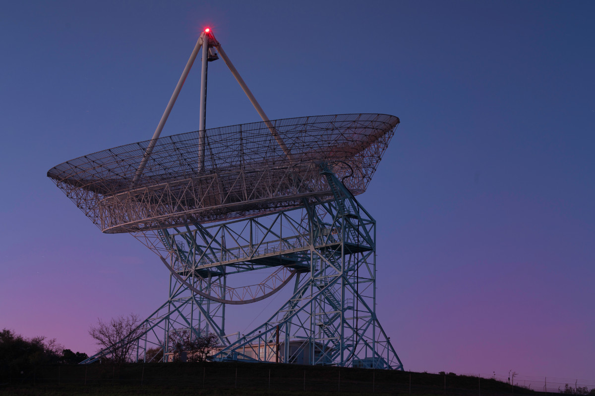 The Stanford Dish, a large radio telescope, in the early morning light. The sky is purple and blue, and the dish is a white metal-lattice structure glowing softly pink in the light. At the top of the dish is a red light. 