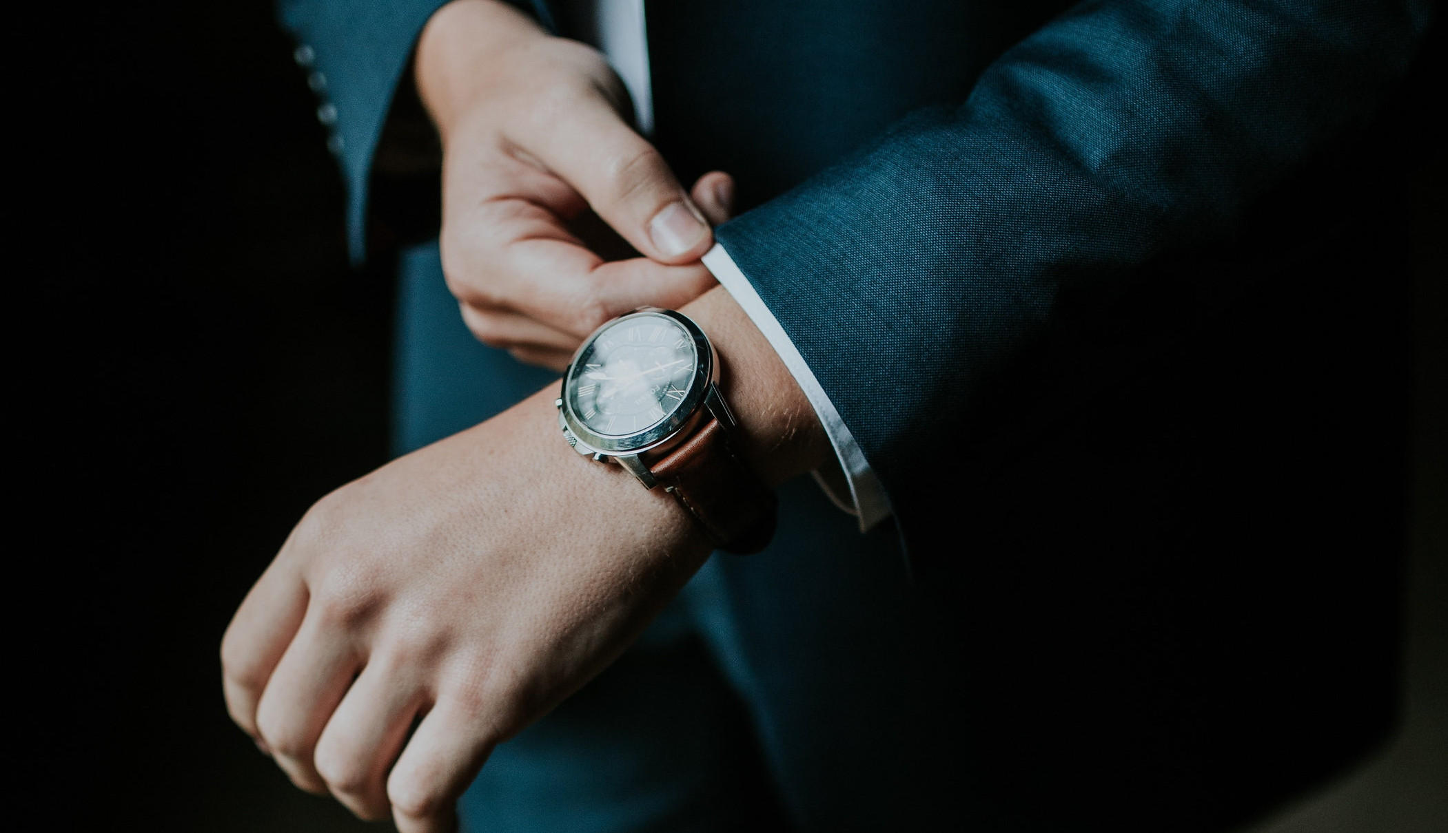 A closeup of a watch on a man's wrist. The man is wearing a nice suite and is pulling on the cuff next to the watch. 