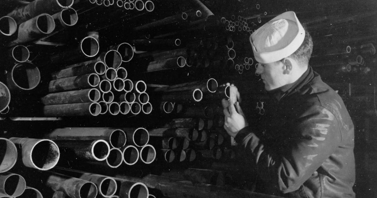 A black and white photo of a man wearing a naval hat filing the ends of metal pipes. 
