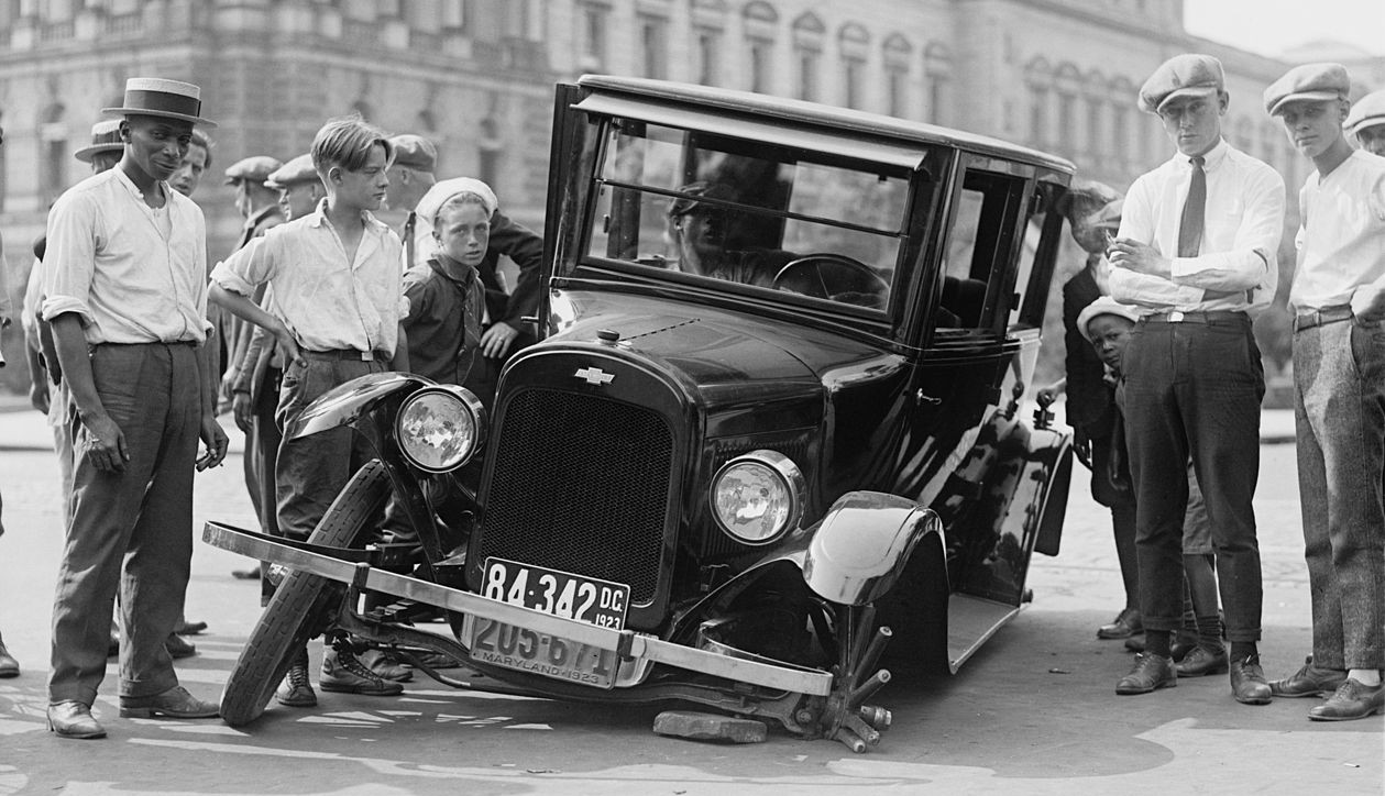 A black and white photo of about a dozen men and boys standing around a broken car taken in Washington D.C. in 1923. One of the car's wheels has splintered and the car is tilted over.