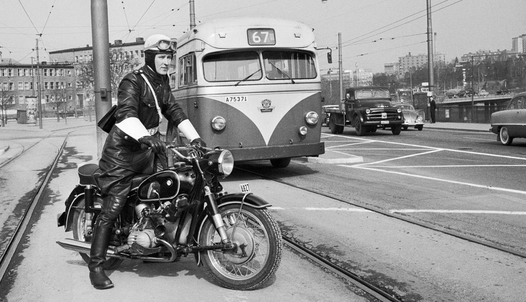 A black and white photo from 1959 of a policeman in full-leather protective gear. He stands over a motorcycle on side of a street in Stockholm as traffic passes by. 