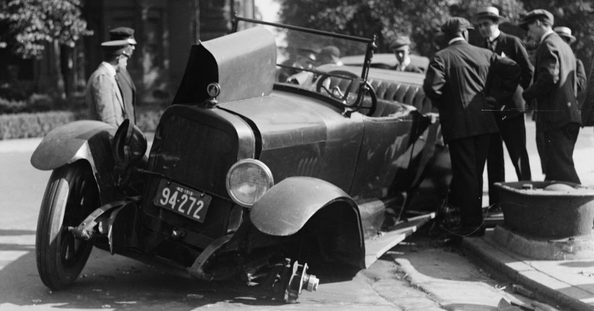 Black and white photo from around 1920 of men in suits with hats crowded around a car that has driven over a curb and lost its front left wheel. 